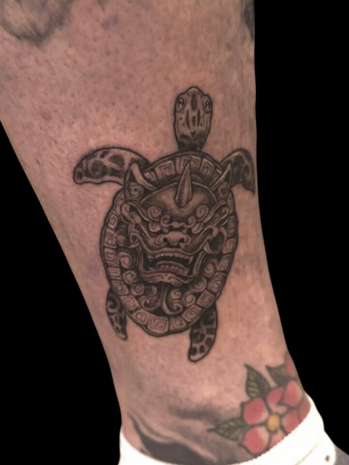 Small turtle tattoo with a carved mask shell above the ankle