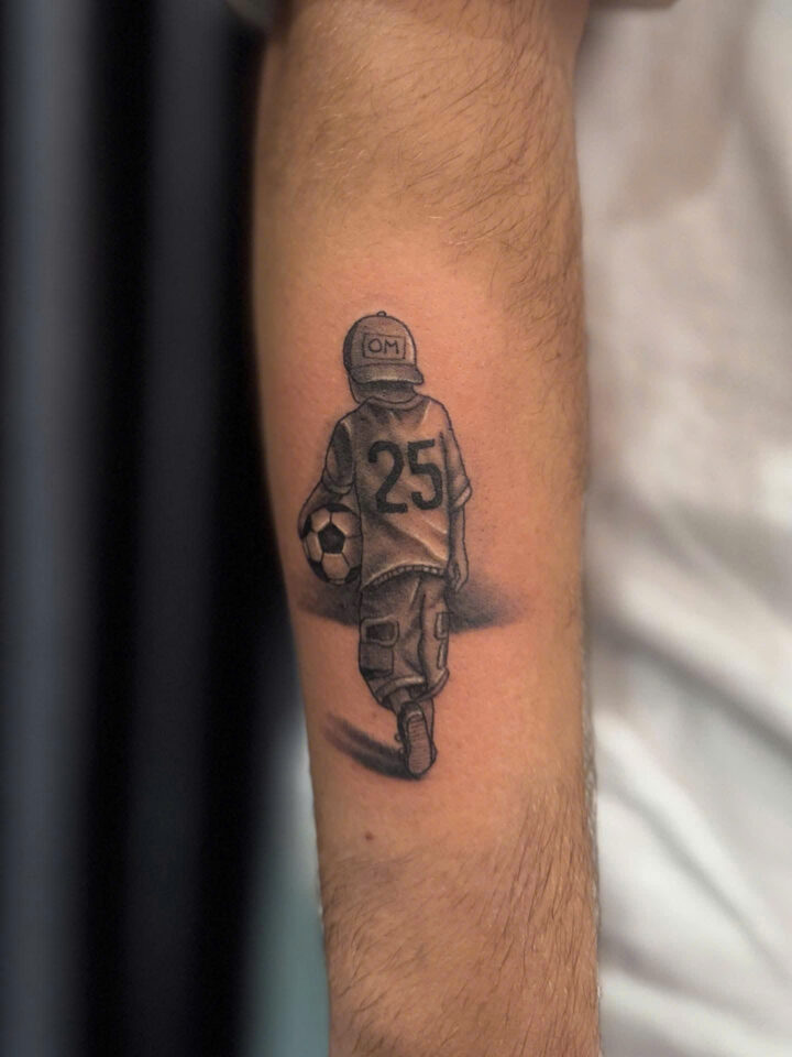 Black and grey tattoo of a young footballer in a number 25 shirt on a forearm
