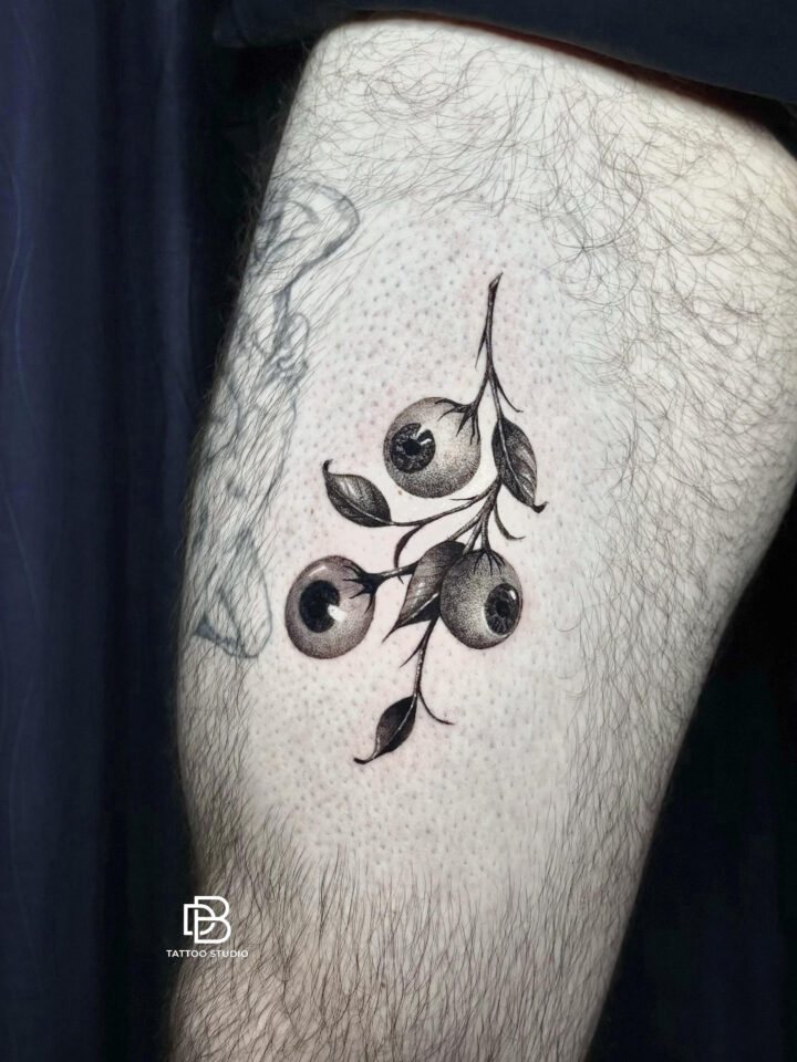 Black and grey berry branch with eyes in place of the fruit, tattooed on a thigh