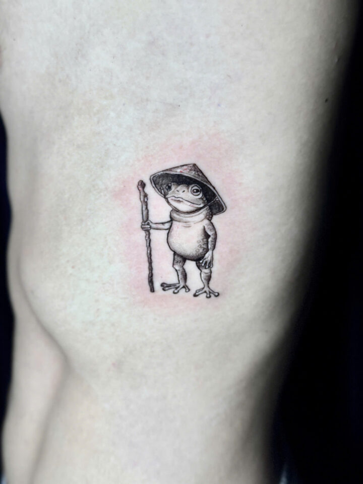 Black and grey frog in a non la hat holding a staff, tattooed on the ribs