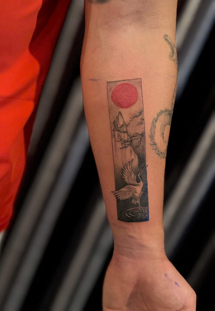 Framed panel tattoo with a red sun over a Vietnamese landscape on a forearm