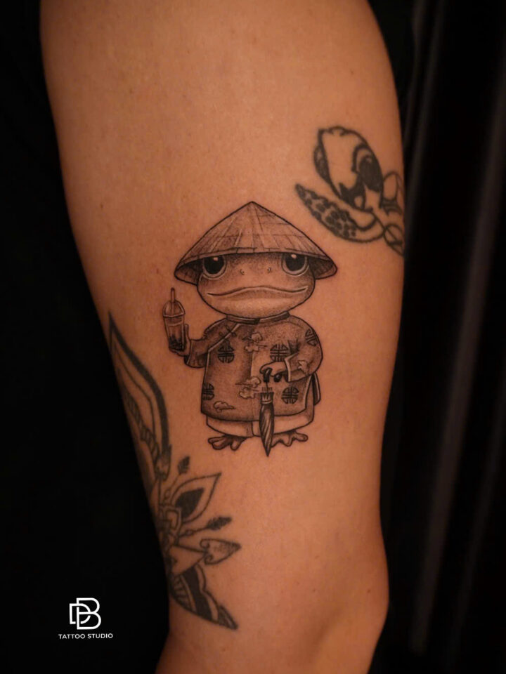 Black and grey tattoo of a frog dressed in a non la and Vietnamese tunic