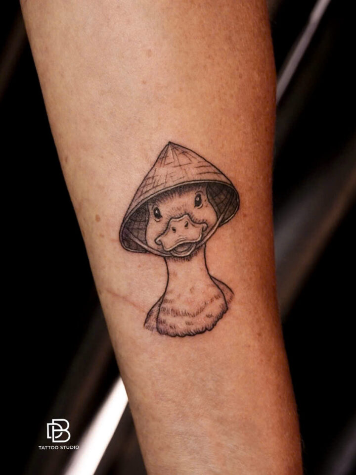 Black and grey duck wearing a non la conical hat, tattooed on a forearm